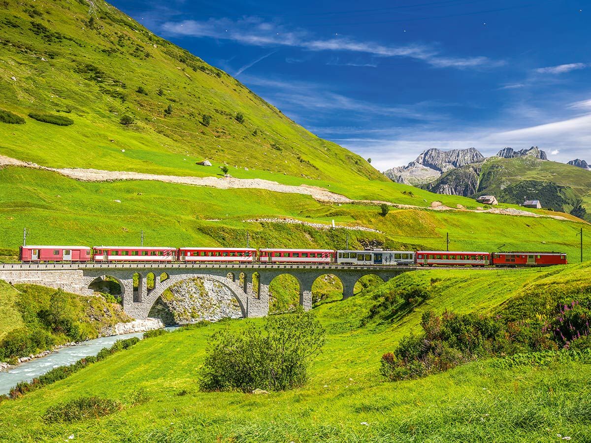 Roter Zug fährt über eine historische Steinbrücke in einer malerischen, grünen Alpenlandschaft mit Fluss und Bergen.