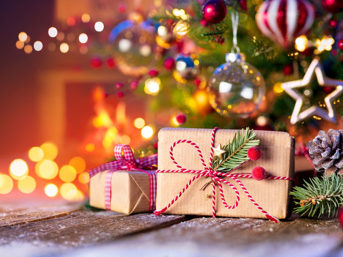 Zwei liebevoll verpackte Weihnachtsgeschenke mit Tannenzweigen und Beeren auf Holztisch vor leuchtendem Weihnachtsbaum.