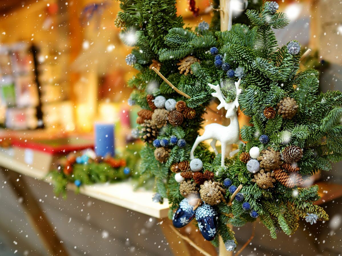 Ein festlicher Weihnachtskranz mit grünem Tannengrün, Tannenzapfen, blauen Beeren, silbernen Kugeln und einem weißen Hirsch vor verschwommenem Weihnachtsmarkt und fallendem Schnee.