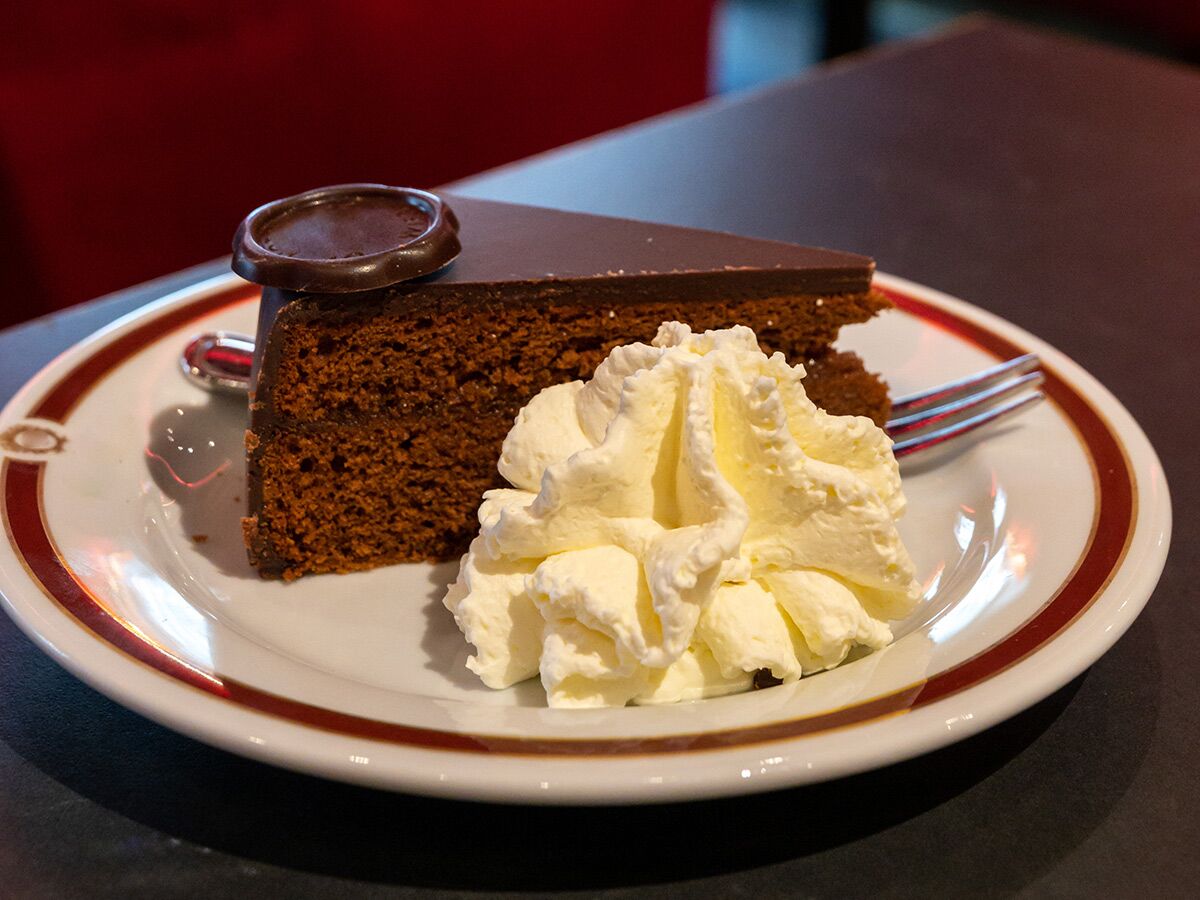 Ein Stück klassische Sacher Torte mit Schokoladenglasur, garniert mit einem Klecks frischem Schlagobers auf einem weißen Teller.