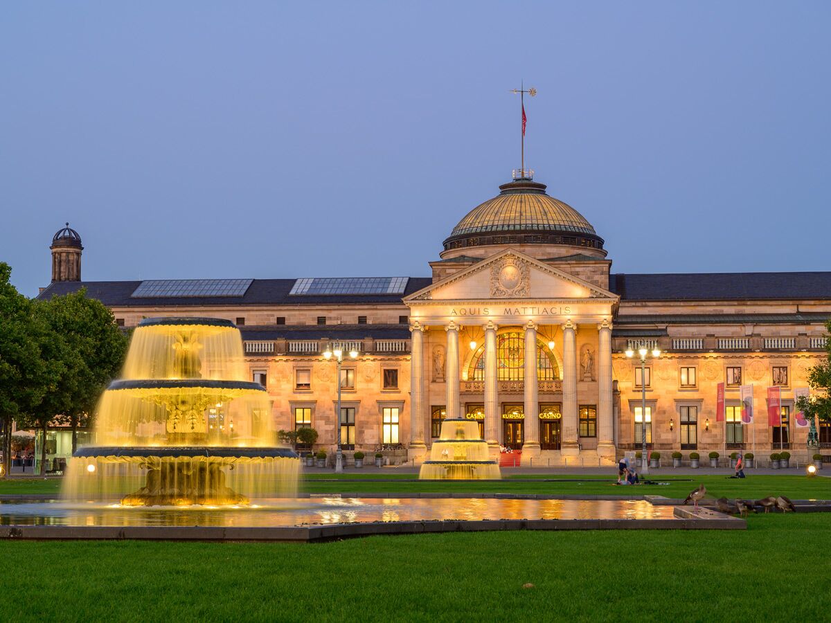 Beleuchtetes Kurhaus Wiesbaden mit großem Brunnen und Grünfläche bei Dämmerung, Menschen entspannen im Vordergrund.