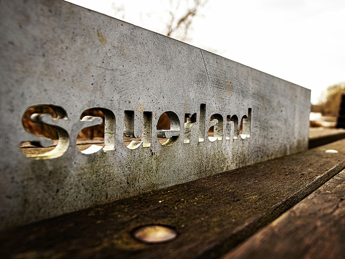 Nahes Bild eines grauen Beton-Schildes mit ausgeschnittenem „Sauerland“-Schriftzug, platziert auf einer rustikalen Holzbank im Freien.
