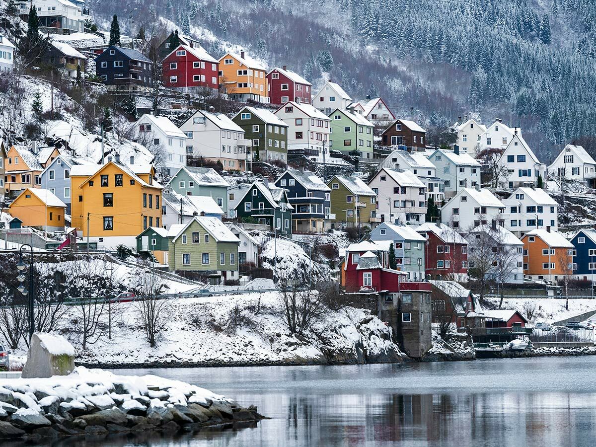 Winterlandschaft mit farbenfrohen Häusern auf schneebedeckten Hängen und einem Gewässer im Vordergrund.