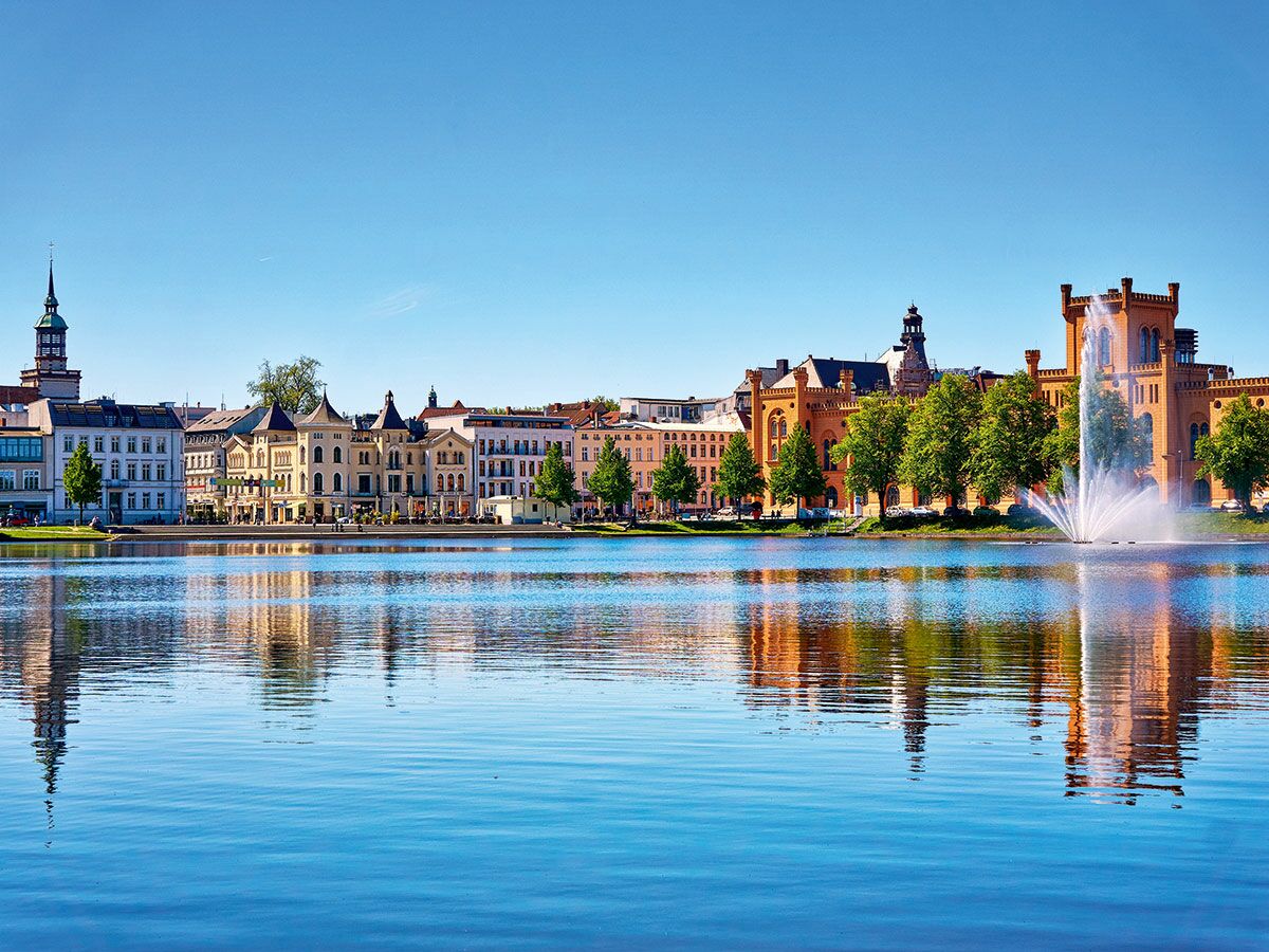 Panoramablick auf Schwerin am Pfaffenteich See mit historischer Architektur, Staatskanzlei und Springbrunnen bei blauem Himmel.