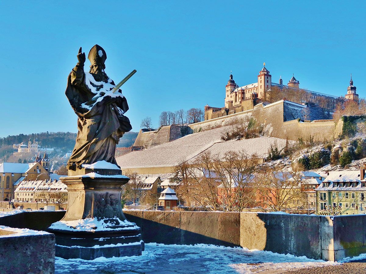 Schneebedeckte Statue auf der Alten Mainbrücke in Würzburg mit der Festung Marienberg und der verschneiten Stadt im Hintergrund.