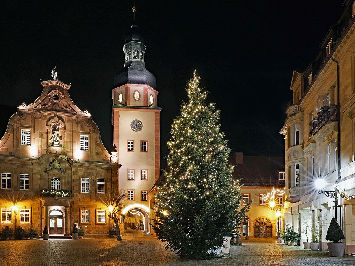 Ein festlich beleuchteter Weihnachtsbaum steht bei Nacht auf einem historischen Marktplatz mit alten Gebäuden und einem Kirchturm in Deutschland.