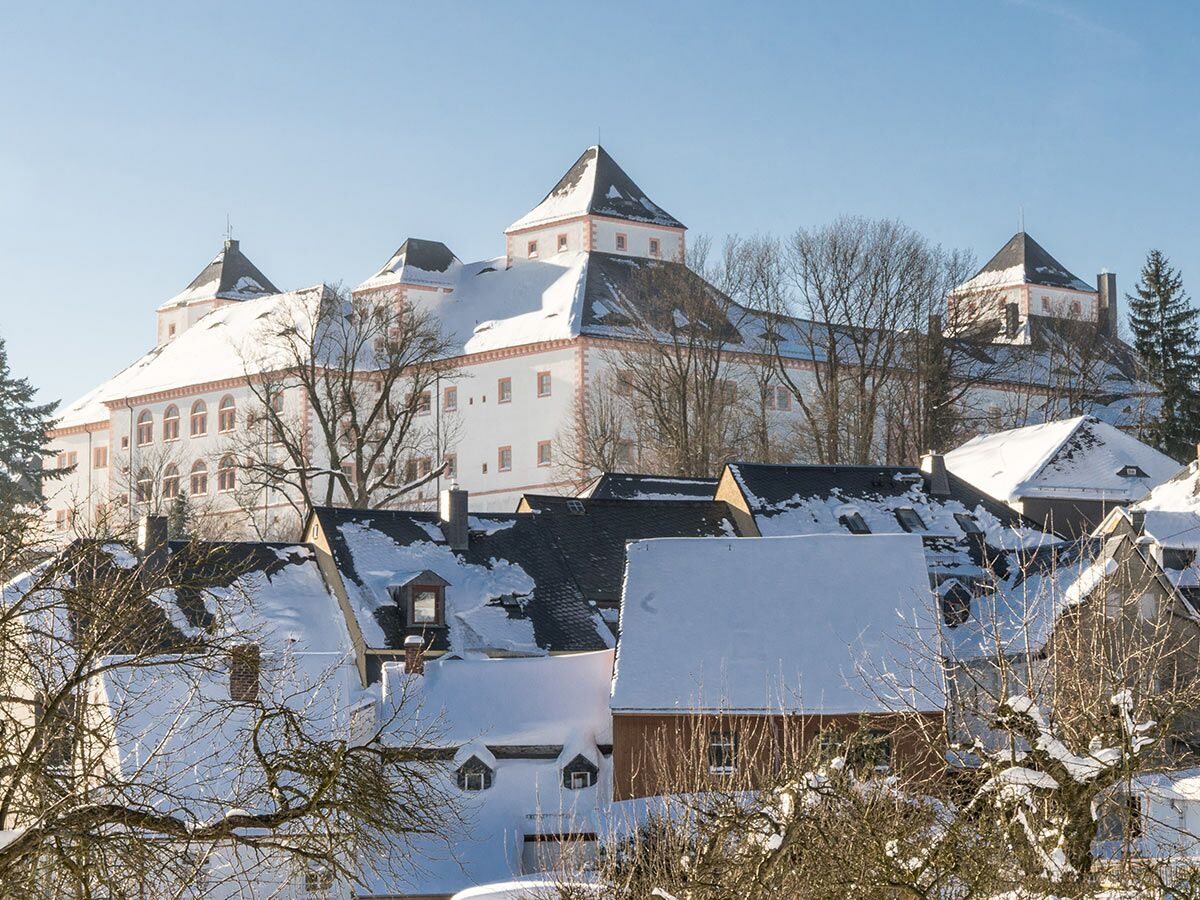 Majestätisches Schloss mit schneebedeckten Dächern und Türmen über einem winterlichen Dorf unter klarem blauem Himmel.