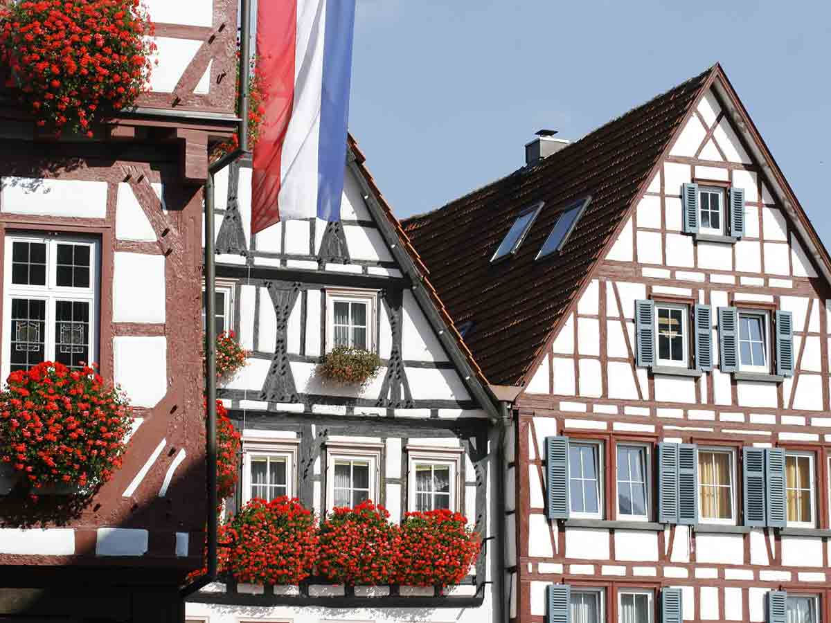 Detailansicht traditioneller Fachwerkhäuser mit roten Geranien in Blumenkästen und einer rot-weiß-blauen Flagge unter blauem Himmel.