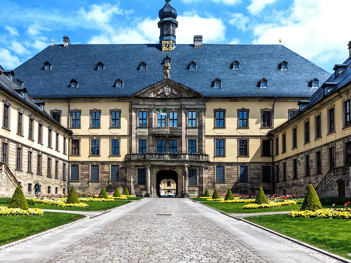 Großes Barockschloss mit gelber Fassade, blauem Dach, Uhrturm, Garten und gepflastertem Weg unter blauem Himmel.