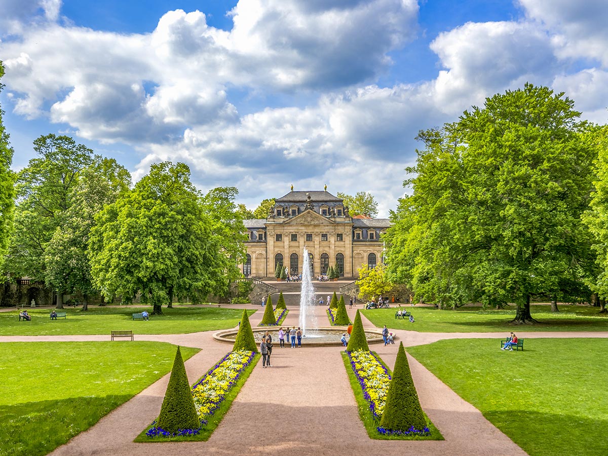 Prächtiger Schlossgarten mit barockem Schloss, großer Brunnenanlage, grünen Bäumen und Blumenbeeten unter blauem Himmel.