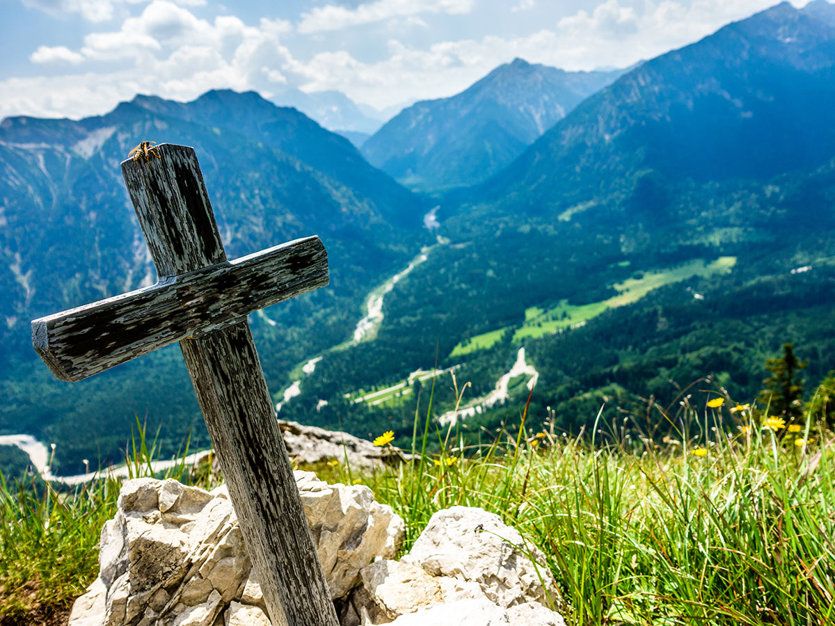 Altes Holzkreuz auf Felsen mit Blick auf ein weites grünes Tal, Flüsse und majestätische Alpen unter blauem Himmel.