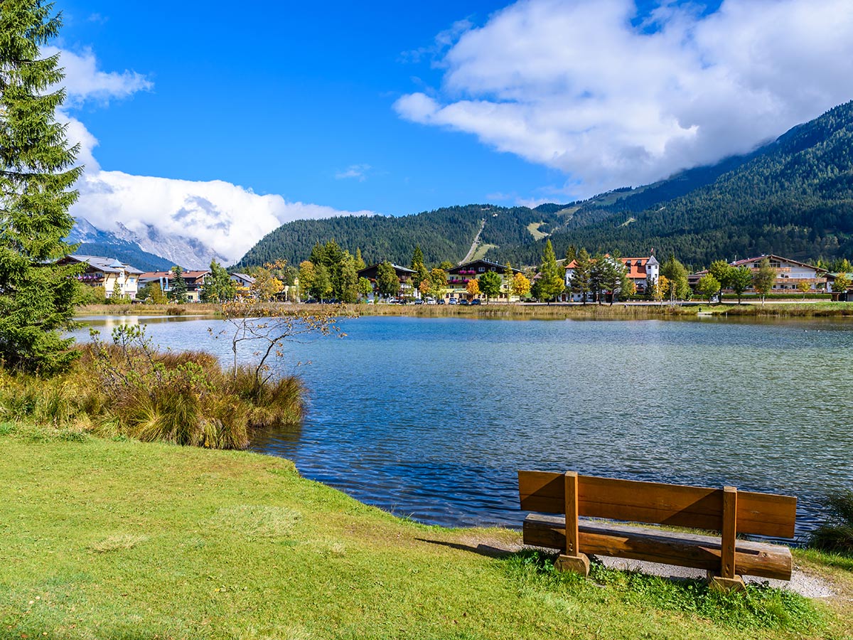 Panoramablick auf einen idyllischen Alpen See mit blauem Himmel, bewaldeten Bergen, einem Dorf am Ufer und einer Holzbank im Vordergrund im Herbst.