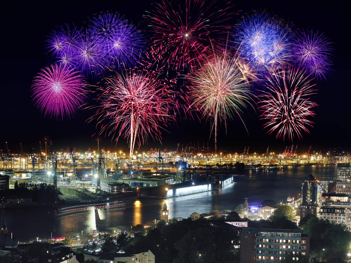 Spektakuläres Feuerwerk über dem Hamburger Hafen bei Nacht, mit bunten Explosionen und beleuchteten Kränen am Wasser.