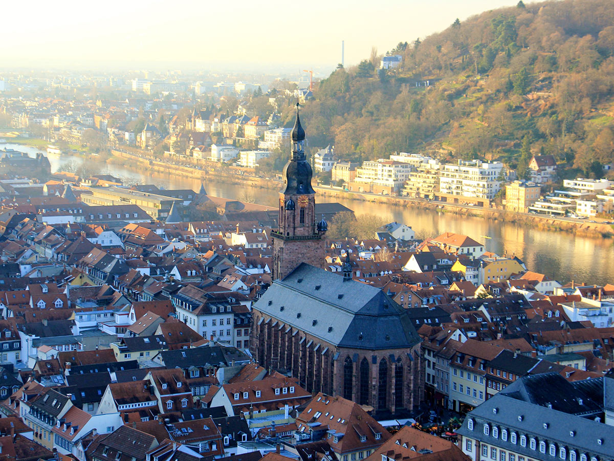 Luftaufnahme der Heidelberger Altstadt mit Heiliggeistkirche, roten Dächern und Neckar bei Sonnenlicht.