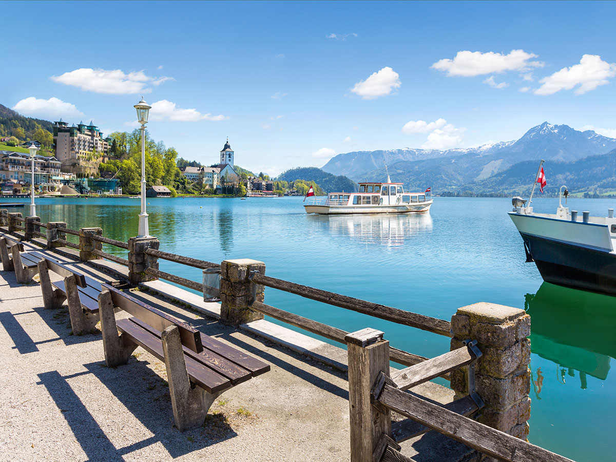 Malerischer Wolfgangsee in Österreich mit Dorf, Kirche, schneebedeckten Bergen und zwei Ausflugsschiffen auf ruhigem Wasser.
