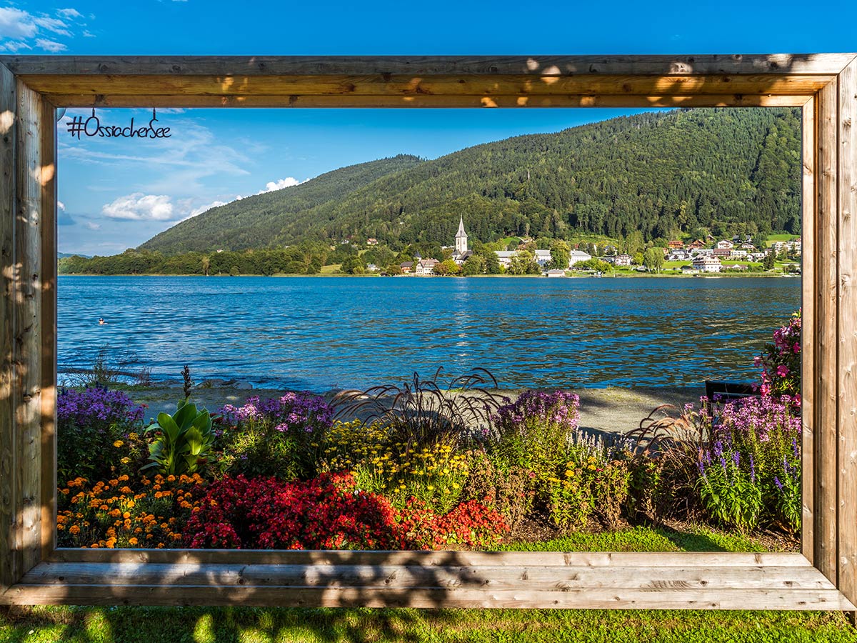 Panoramablick auf den Ossiacher See mit Bergdorf, gerahmt von bunten Blumen und Holzsteg. #Ossiachersee