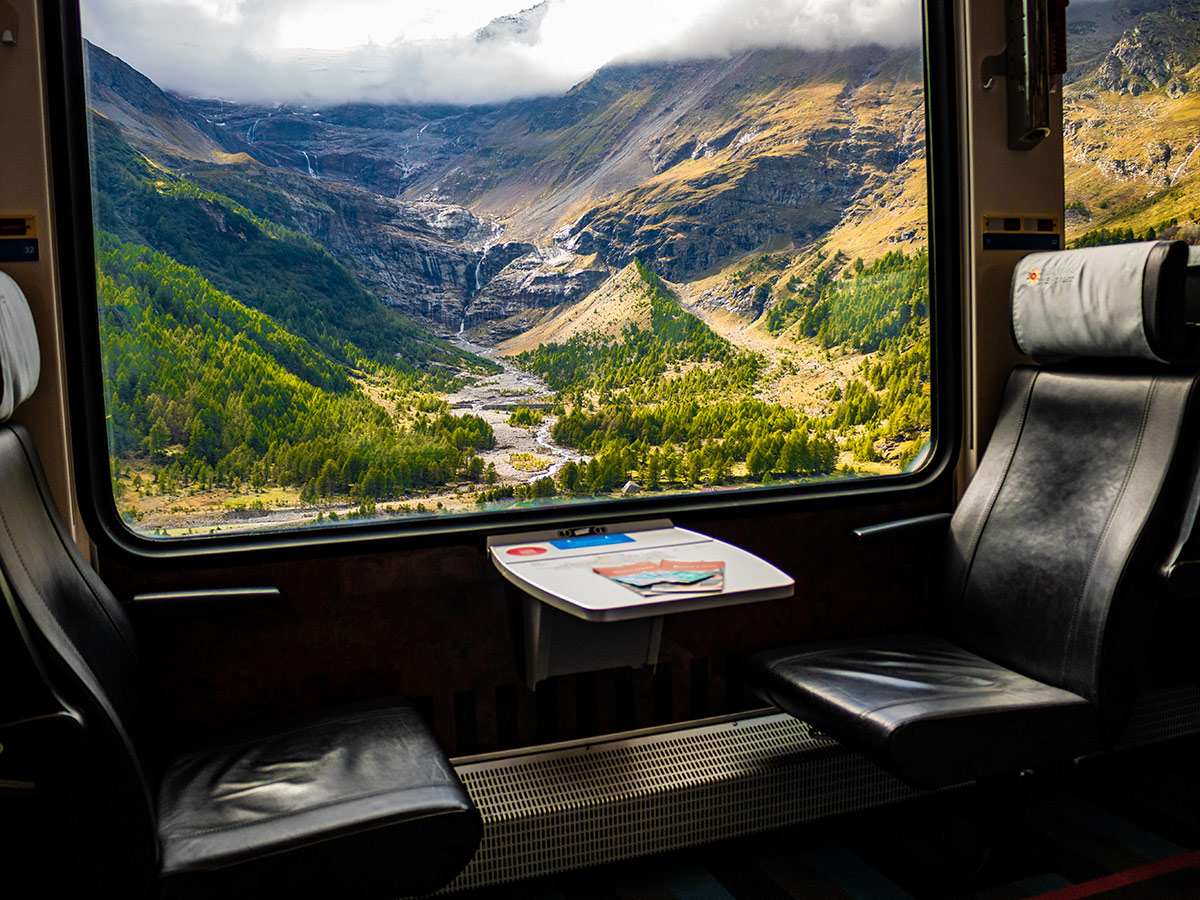 Panoramablick aus Zugfenster auf grüne Alpenlandschaft mit Tal, Fluss, Wäldern und Wasserfällen unter bewölktem Himmel. Leere Ledersitze.